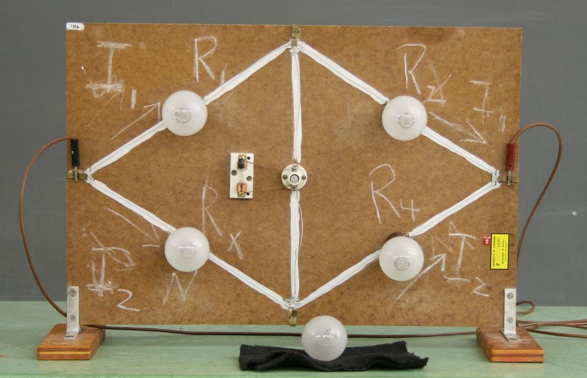 Wheatstone bridge board with five light bulb sockets in a diamond pattern connected by white conducting strips on brown fiberboard, chalk labels R1 through R4 and Rx, a small knife switch at center, power connections on the sides, mounted on wooden feet. A spare bulb sits on the bench below.