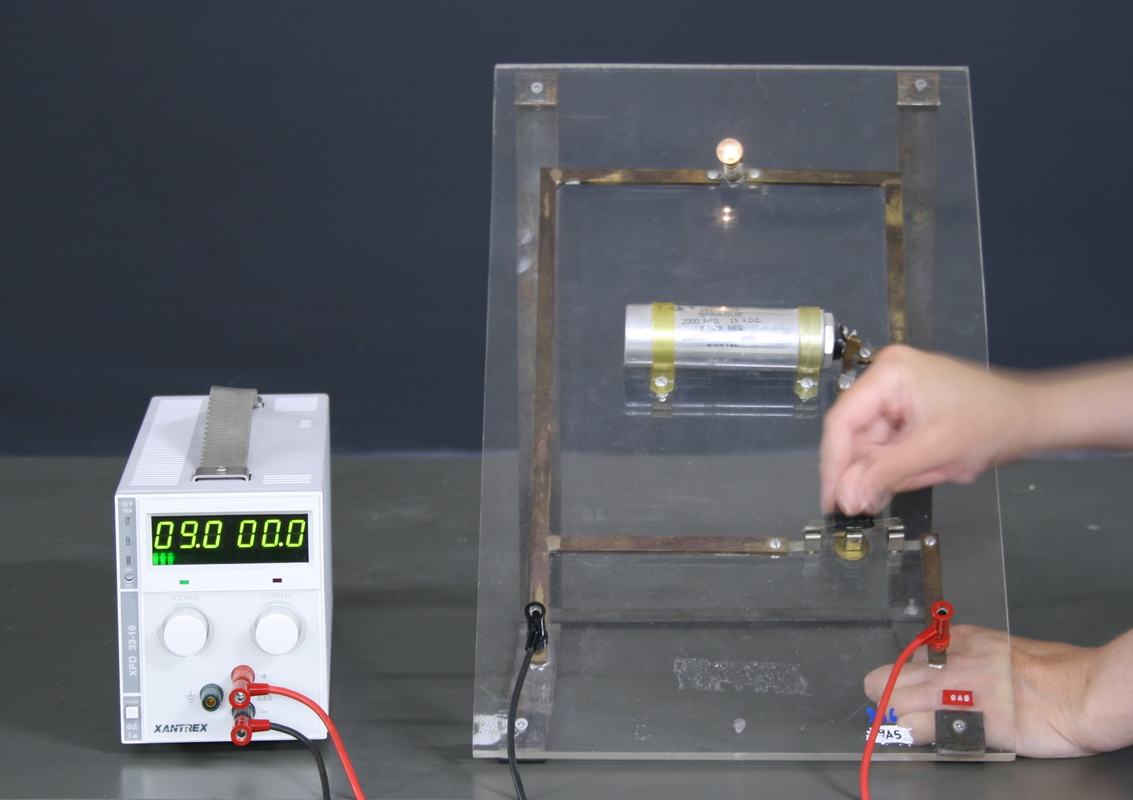 Wooden-framed RC demonstrator board with transparent acrylic panel, large horizontal electrolytic capacitor, small incandescent bulb at top, and knife switch at bottom right, connected to a Xantrex DC power supply reading 9.0V
