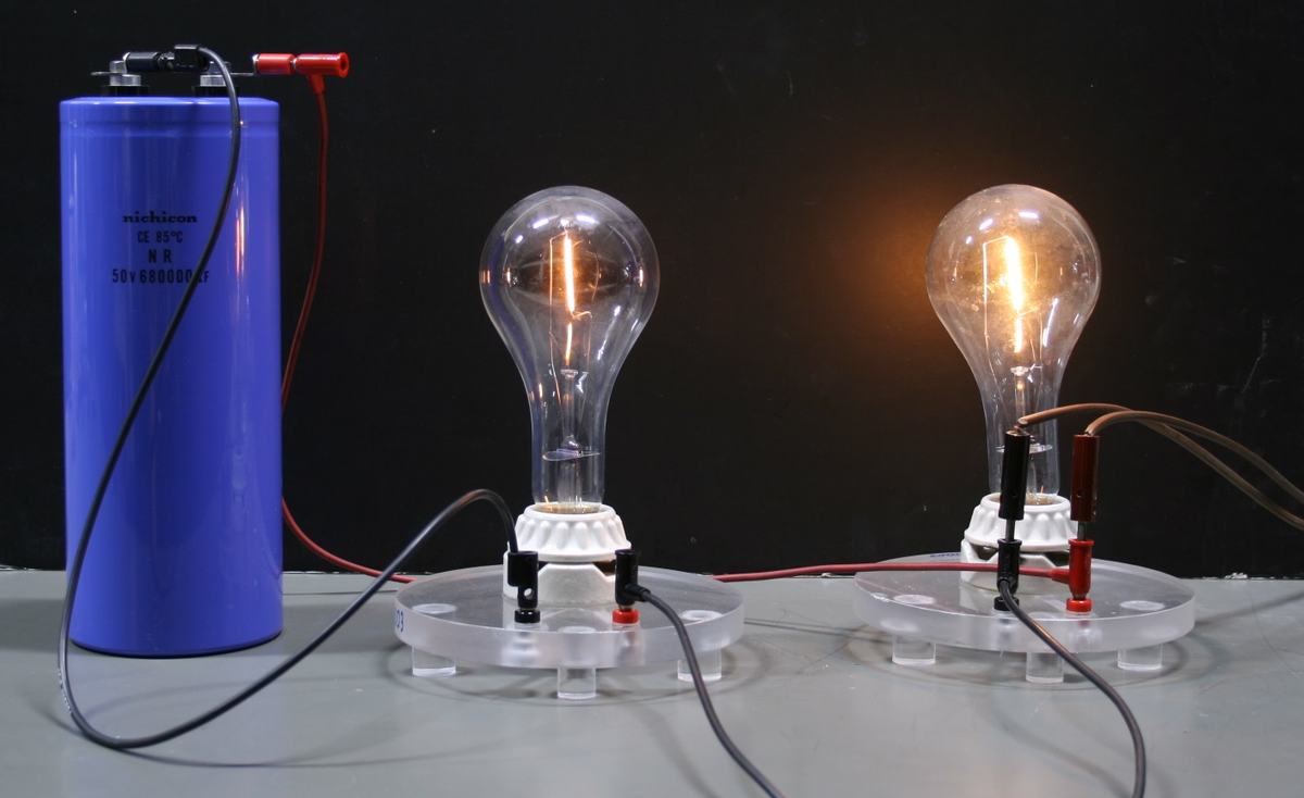 Two incandescent bulbs on clear plastic bases with white ceramic sockets connected by wires to a large blue Nichicon 50V capacitor; the left bulb near the capacitor is dim while the right bulb is brightly lit
