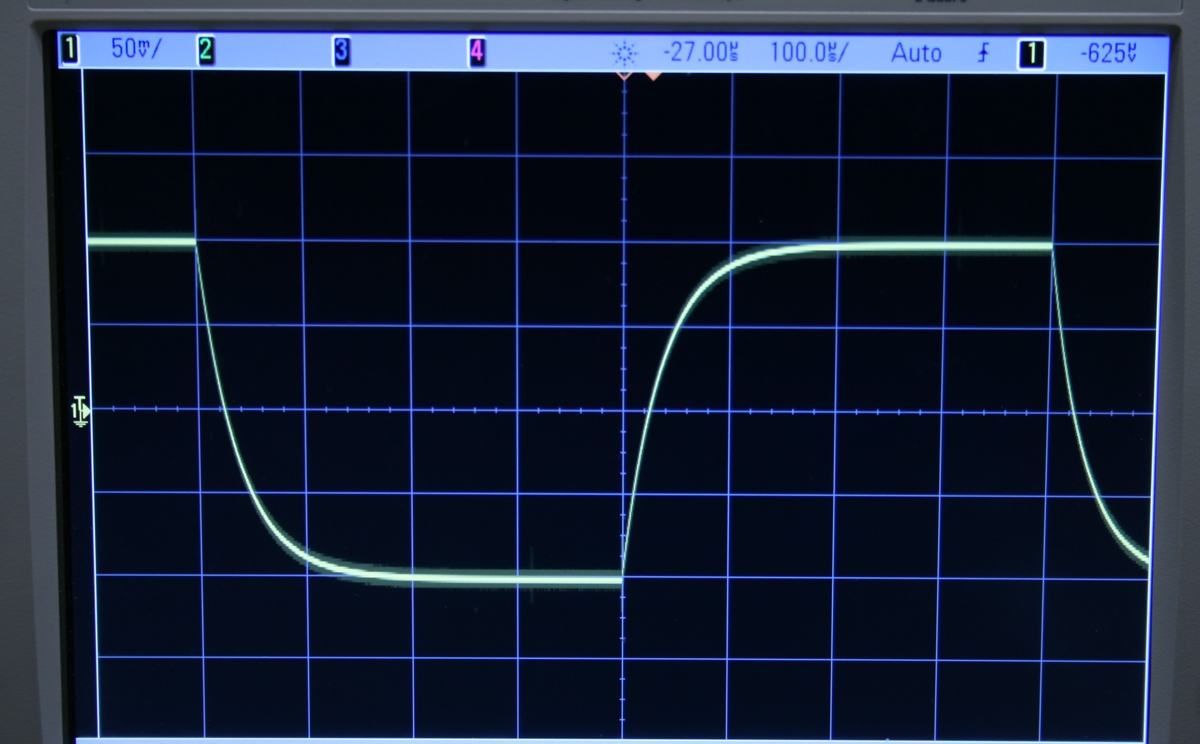 Oscilloscope close-up showing two traces: square wave input (top) and capacitor voltage with exponential rise and fall (bottom), displaying a full charge/discharge cycle
