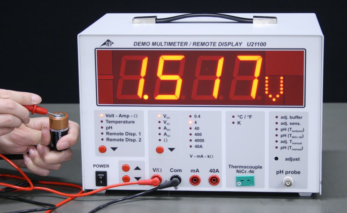 Front panel of 3W Demo Multimeter U21100 with large red LED display reading 1.517 V; hand holds a battery against the test leads