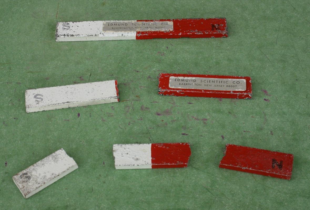 Several broken bar magnet pieces (red-painted and bare metal) with Edmund Scientific labels on a green surface