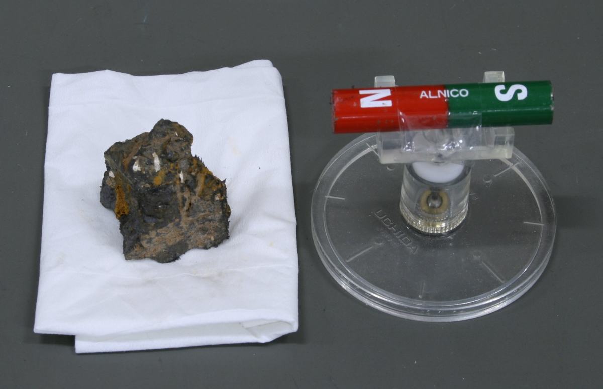 Lodestone on white cloth next to an ALNICO bar magnet (red N, green S) on a clear plastic pivot stand