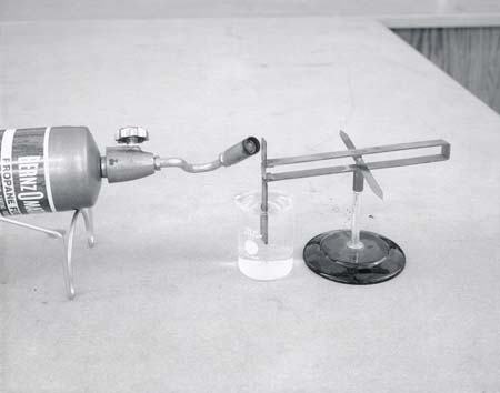 Bernzomatic propane torch on stand heating a copper-constantan thermocouple junction, with compass needle on stand nearby and small beaker of ice water