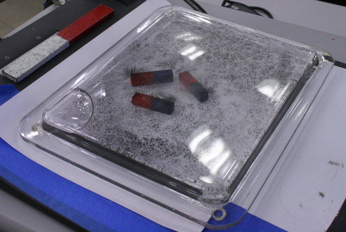 Magnetic field projectual (sealed clear plastic tray with iron filings and glass stirring rod) resting on a surface with two bar magnets underneath showing field patterns