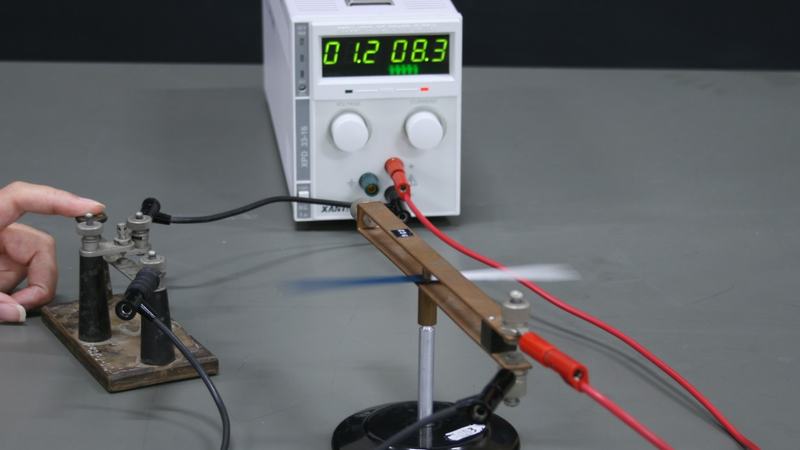 Oersted's experiment apparatus: copper loop on a small stand with compass needle inside, connected via black and red leads to a white DC power supply reading 01.2 V and 08.3 A, with a terminal block on the left and a hand adjusting it