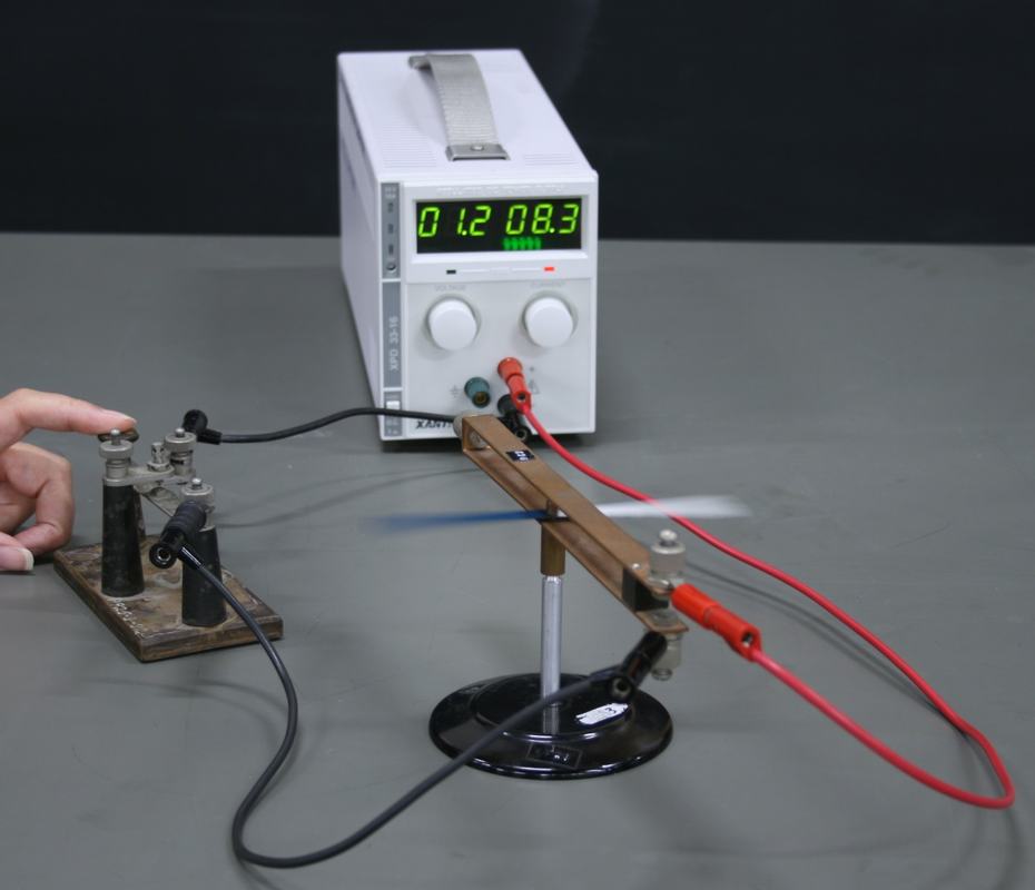 Oersted's experiment apparatus: copper loop on a small stand with compass needle inside, connected via black and red leads to a white DC power supply reading 01.2 V and 08.3 A, with a terminal block on the left and a hand adjusting it