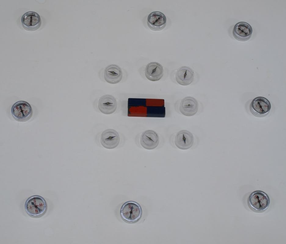 Thirteen compasses in a grid pattern around a single small red-and-blue bar magnet in the center on a white surface