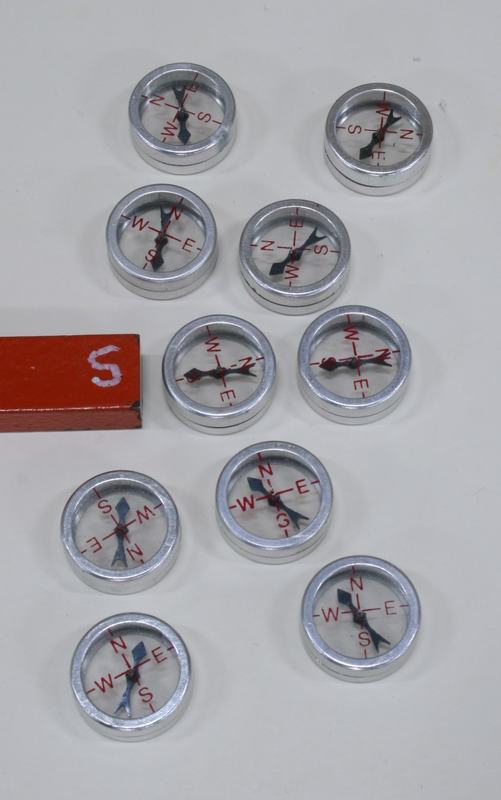 Ten silver-cased compasses clustered near the red S-pole end of a bar magnet on a white surface