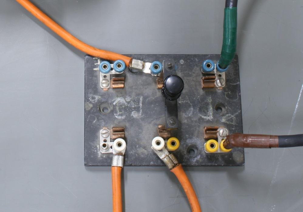 Close-up overhead view of double-pole switch junction box with six brass terminal posts, orange and green insulated wires connected, black and brown wires, metal plate base