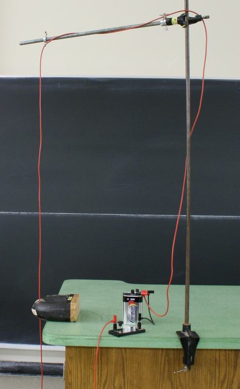 Clamp stand on lecture bench with red wire hanging from horizontal rod, radar magnet on its side at edge of bench, Energizer battery and telegraph switch connected via red wires