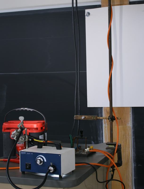 Side view showing the fiber optic lamp (blue box), red Optima battery with alligator clips, wooden support rod with bench clamp, white paper backdrop, and orange insulated leads