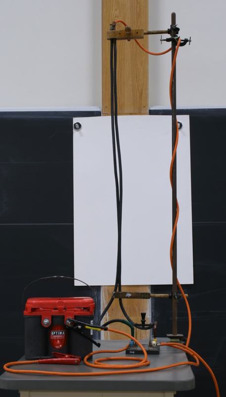Two long black rubber-insulated wires hanging from a wooden clamp on a vertical support rod, with a white paper backdrop behind them, a red Optima 12V battery and switch on the bench below, and heavy orange leads connecting the circuit