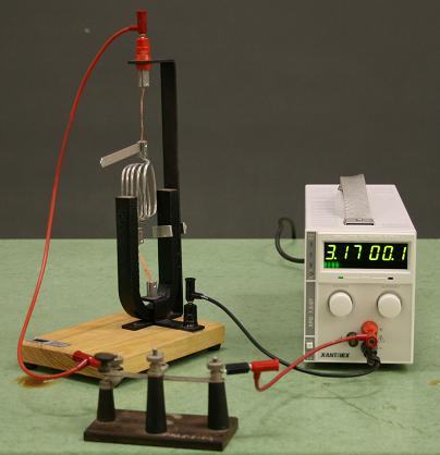 Wire coil suspended between poles of a horseshoe magnet on a wooden base, connected via red wires to a Xantrex DC power supply reading 3.17 V