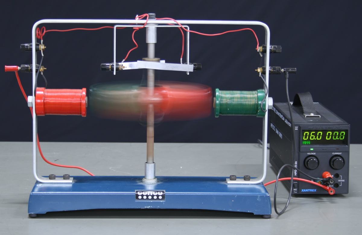 CENCO motor-generator model running as a motor: armature is a blur spinning between red (left) and green (right) field coils, mounted on white metal frame with blue base. Xantrex power supply at right reads 6.0V, 00.8A. Four binding posts with red wires visible on top crossbar.