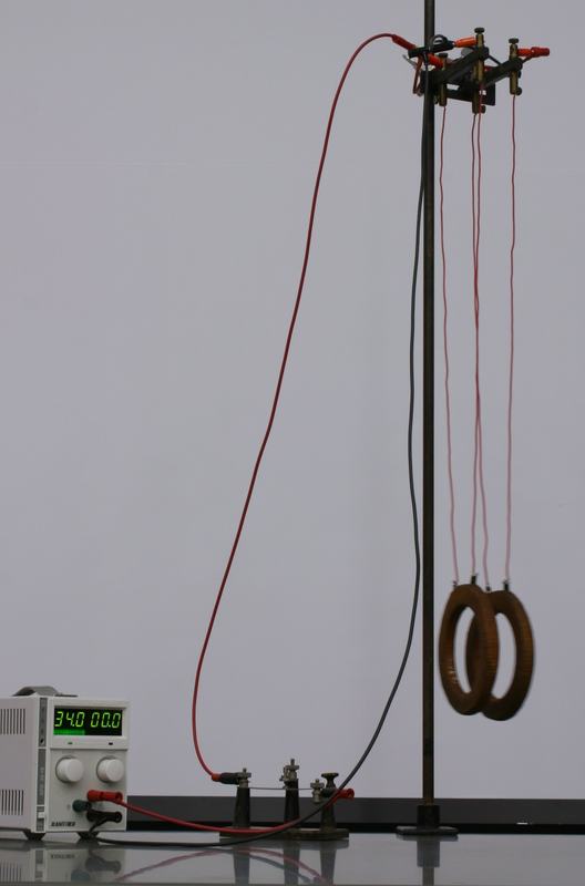 Two copper coils suspended by thin red wires from a support rod on a ring stand, connected via binding posts and red/black leads to a DC power supply reading 34.0V