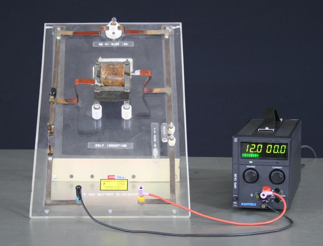 Acrylic self-induction demonstrator board with iron-core inductor, copper bus bars, porcelain standoffs, incandescent bulb at top, and knife switch at lower left, connected to Xantrex HPD 15-20 power supply set to 12.0 V