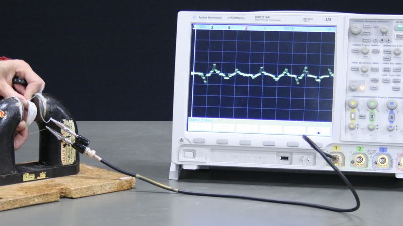 Hand holding a flexible coil between the poles of a large C-shaped permanent magnet on a wooden base, connected via BNC cable to an Agilent oscilloscope displaying induced voltage pulses