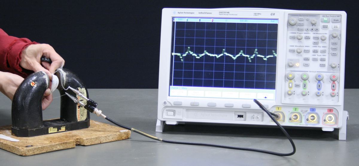 Hand holding a flexible coil between the poles of a large C-shaped permanent magnet on a wooden base, connected via BNC cable to an Agilent oscilloscope displaying induced voltage pulses