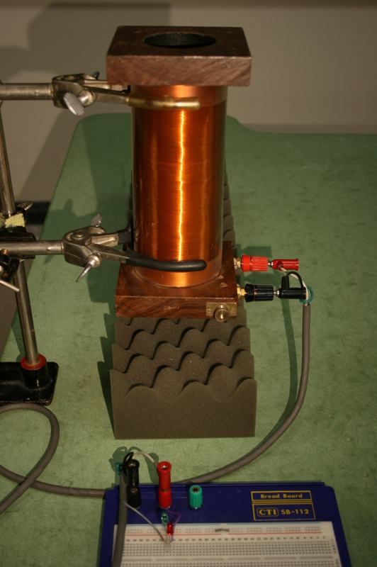 Copper wire induction coil on wooden base with banana jacks, clamped vertically on a stand with eggshell foam underneath, connected by leads to a breadboard with two LEDs