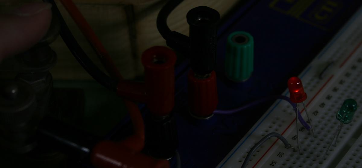 Close-up of breadboard with red LED illuminated and green LED dark, showing banana plug connections