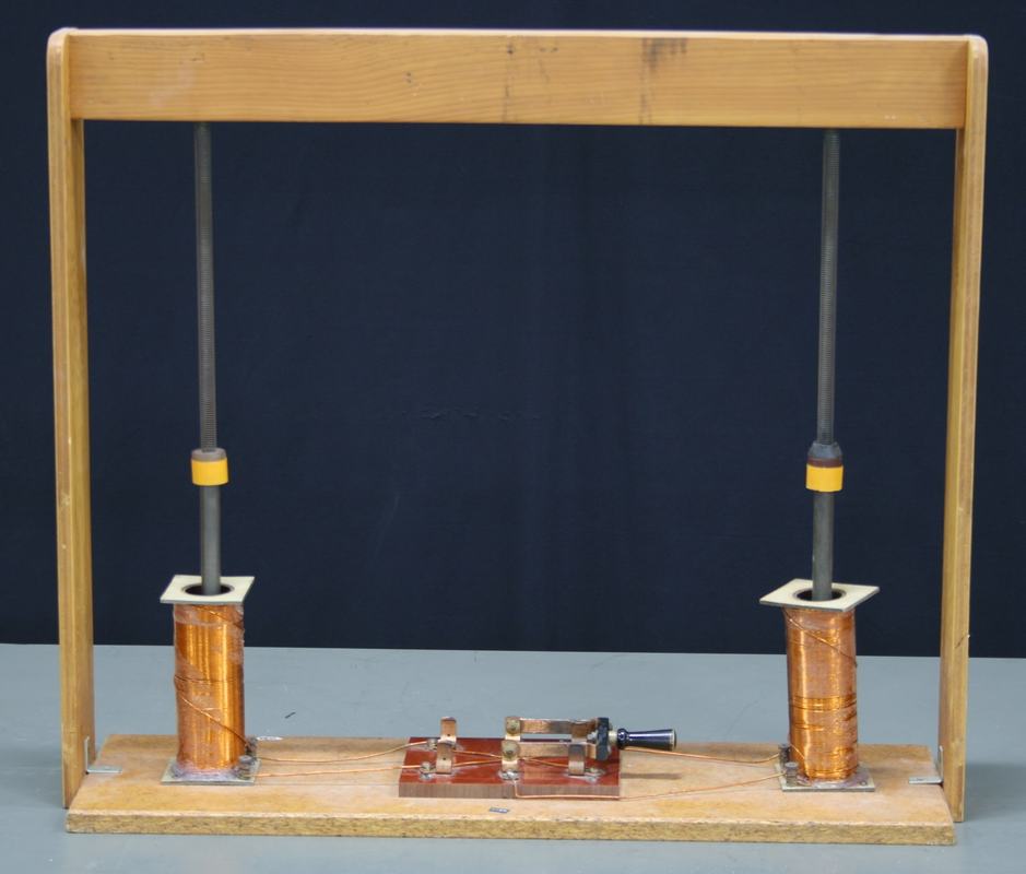 Wooden frame with two vertical rods suspended from springs at the top crossbar, each passing through a copper-wound solenoid coil at the base, yellow position markers on the rods, and a polarity-reversal switch board mounted between the coils