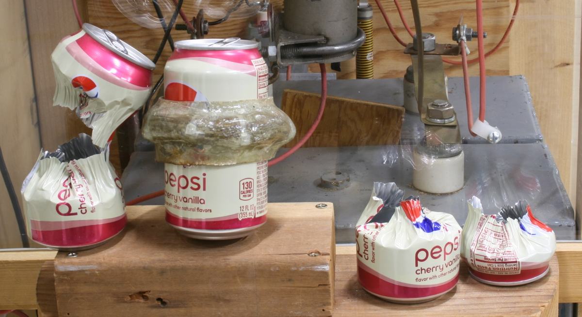Crushing coil wrapped in fiberglass and tape atop the apparatus, surrounded by several crushed Pepsi cans showing various degrees of compression