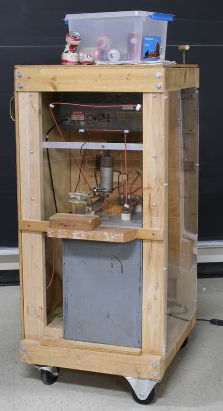 Full can crusher apparatus on rolling cart with wooden frame, clear acrylic safety panels, high-voltage warning labels, and storage bin of soda cans on top