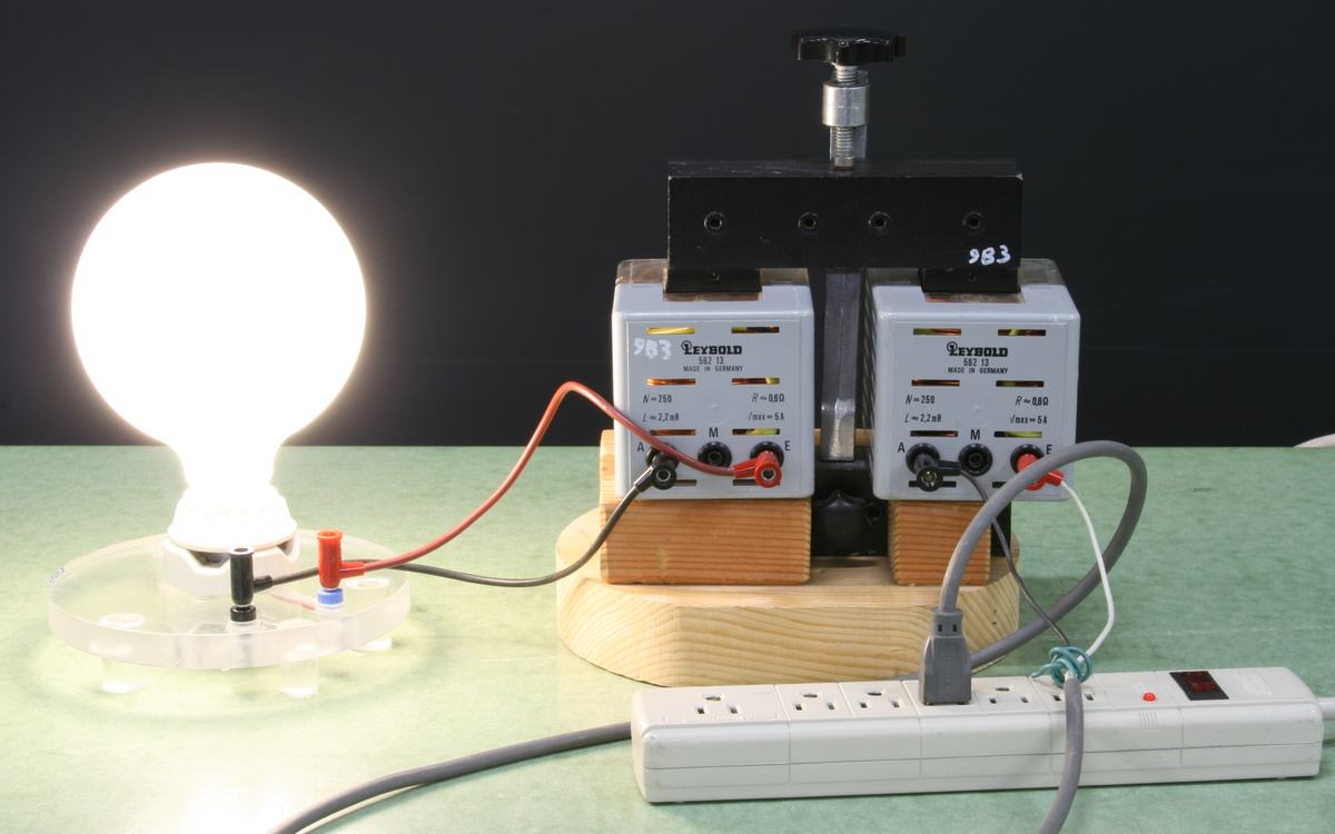 Assembled dissectable transformer on wooden base: black iron core frame with two white Leybold 983 coil units mounted side by side, illuminated round light bulb on left, power strip below