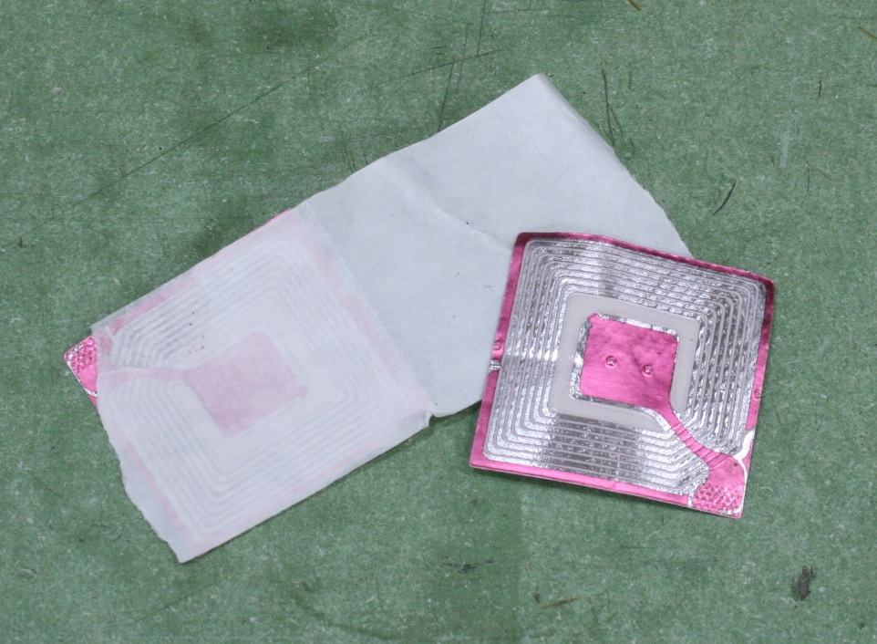 Two anti-theft stickers: one intact showing pink spiral pattern through translucent backing, one peeled open revealing metallic spiral coil antenna and small chip
