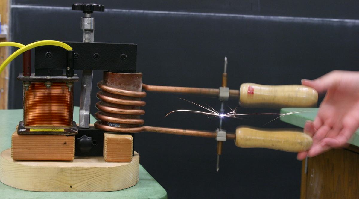 Demountable transformer on wooden base with 250-turn primary coil (left) and 5-turn copper helix secondary coil, connected to wooden-handled nails producing bright sparks at contact
