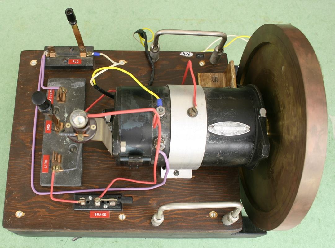 Overhead view of motor-generator board showing switches labeled FLD, GND, and BRAKE, DC motor with bronze flywheel, and colored wiring on wooden base