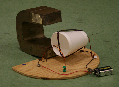 Simple motor: wire coil mounted on a coffee cup with a permanent magnet behind it, on a wooden base with wire contacts for a 9V battery