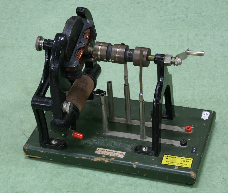 Demonstration AC-DC generator on dark green base with wooden-handled hand crank on left, exposed copper armature windings in center between black field coil poles, commutator and slip rings on the shaft, and terminal posts on the base