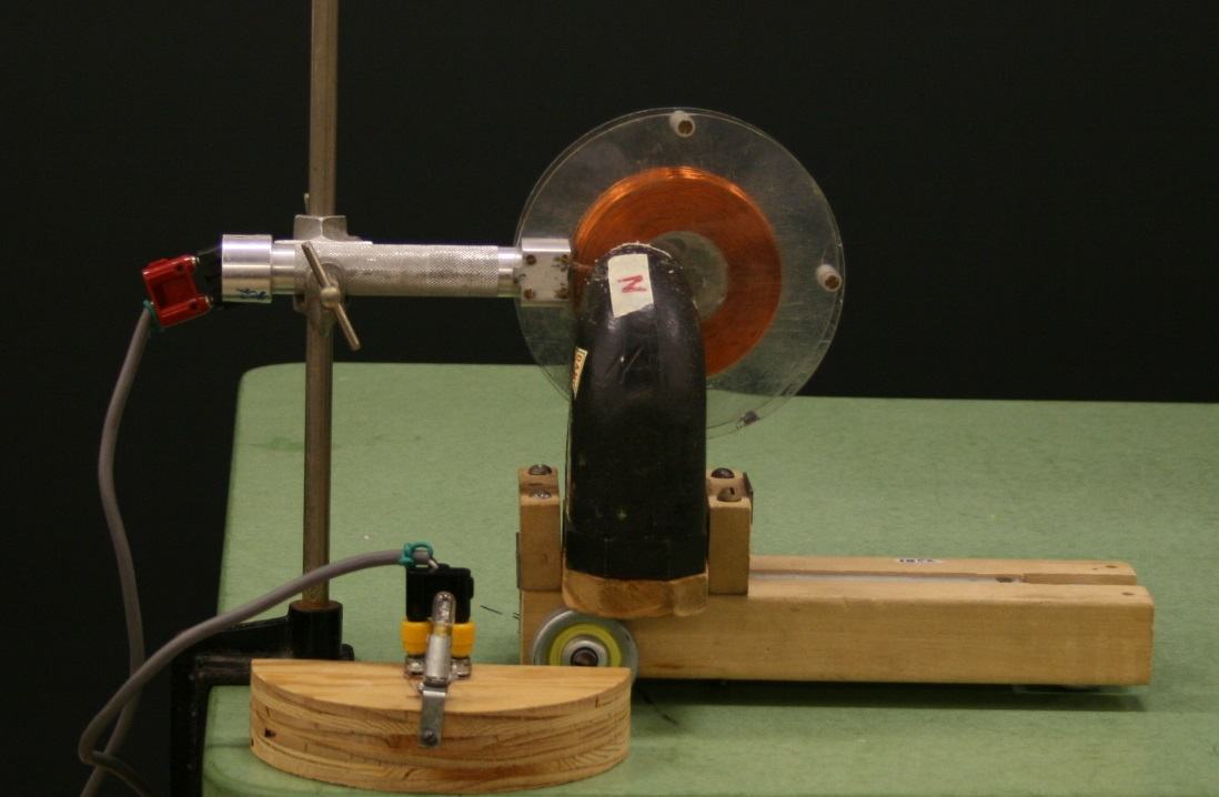 Linear generator apparatus: copper coil with clear plastic guard plates on a metal handle mounted to a clamp stand, positioned between the poles of a large black radar magnet on a wooden cart with wheels. A miniature lamp in a base is connected by wires on the left.