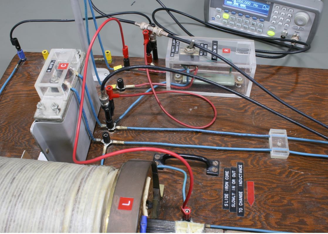 Close-up of LCR board showing transparent component enclosures, binding posts, banana plug wiring, function generator, and wire spool inductor with label "Slide knob over to change inductance"