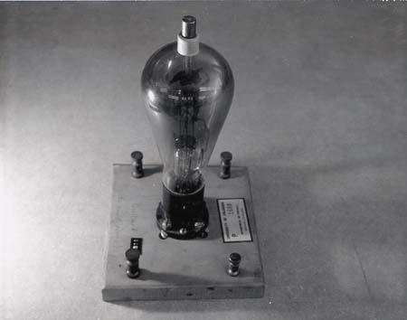 Large vacuum tube rectifier mounted on wooden base with binding posts. Clear glass envelope reveals internal plate structure surrounding the filament. A label is visible on the base.