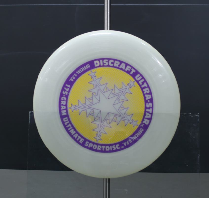 White Discraft Ultra-Star disc with purple and yellow logo, hanging from ring clamp under normal room lighting