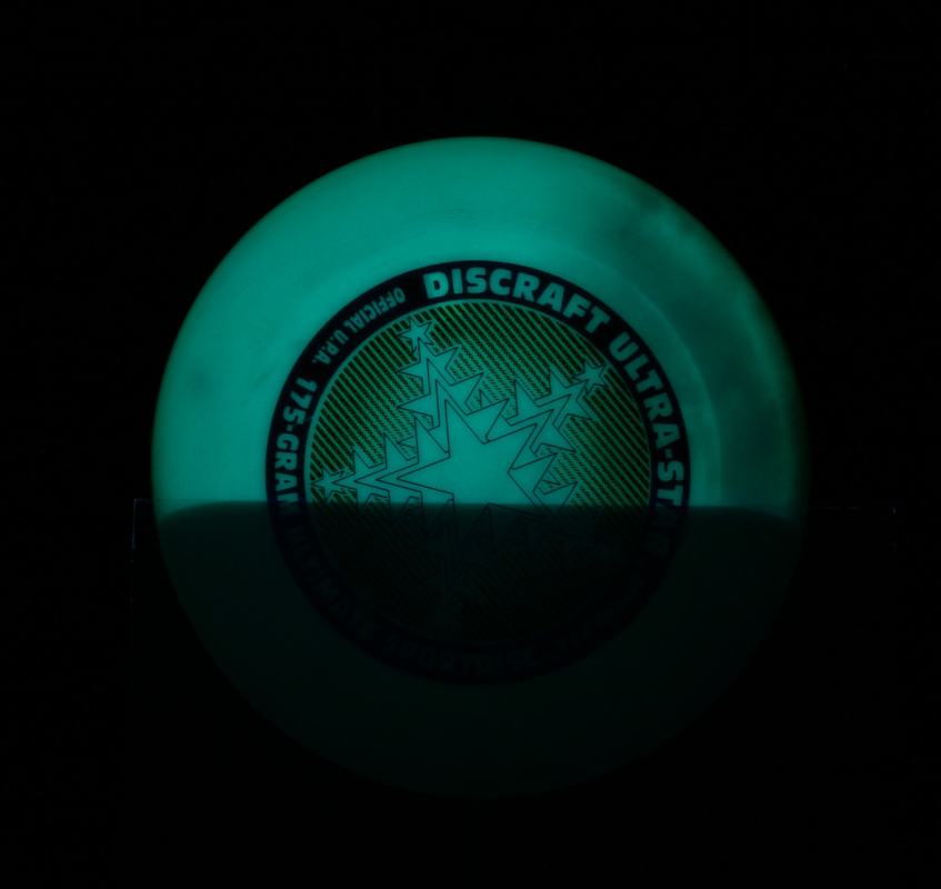 Phosphorescent Discraft Ultra-Star disc glowing green under UV light, partially shadowed by glass plate blocking part of the UV