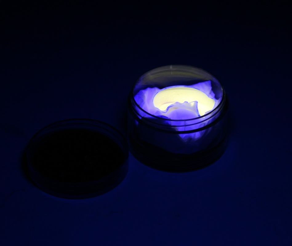 CFL phosphor in open container glowing yellow-white at center with blue-purple edges under shortwave UV