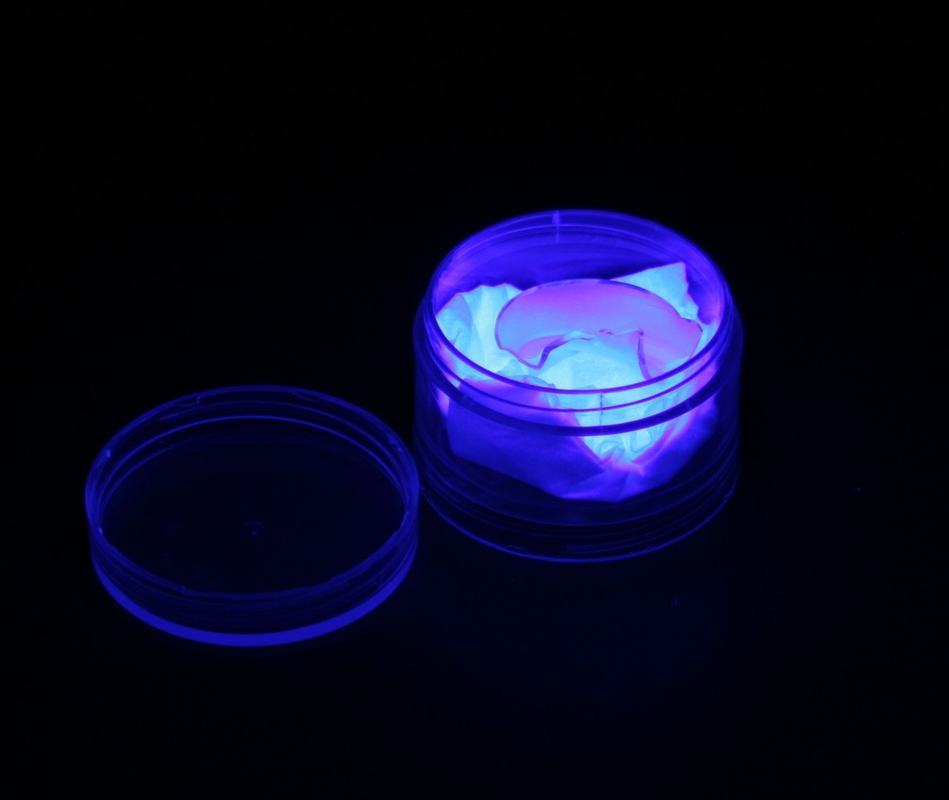 CFL phosphor in open plastic container fluorescing blue-purple under shortwave UV in darkness, lid sitting beside container