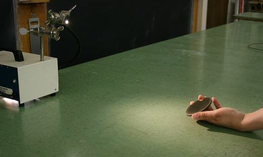 Optical cable light source on a lecture bench with a hand holding a bronze mirror disc, reflecting light toward a wall