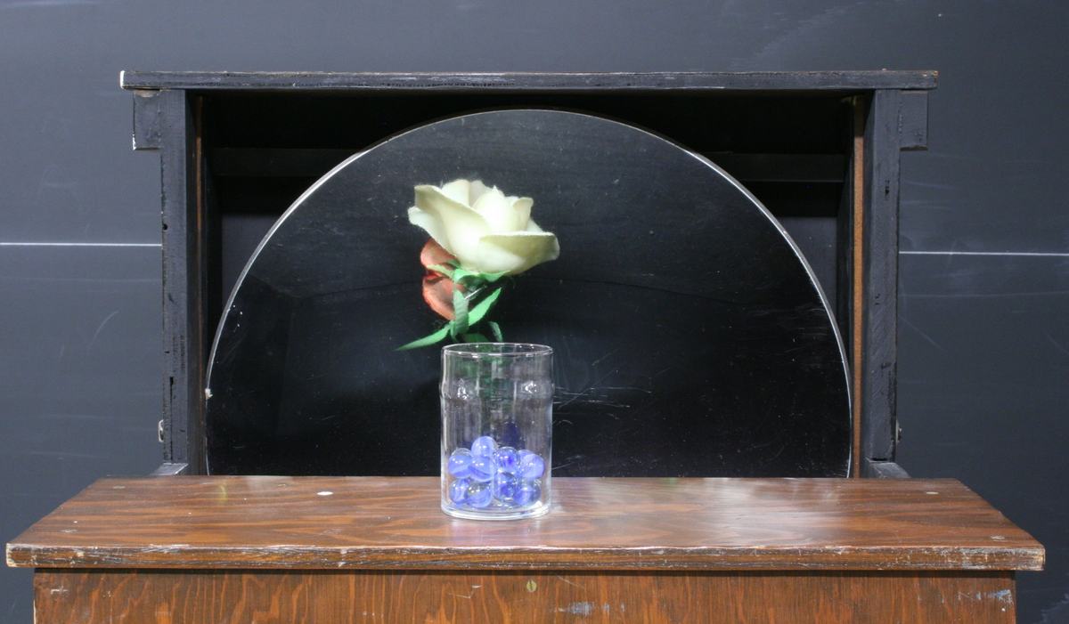 Half-open wooden box with large concave mirror visible through the opening; a white rose appears to stand upright in a clear glass with blue marbles on the wooden lid
