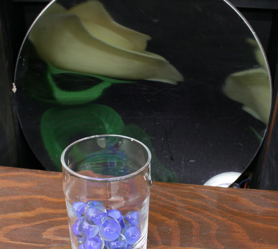 Close-up of the clear glass with blue marbles on the wooden lid, with the large concave mirror behind showing a magnified reflection of the rose image