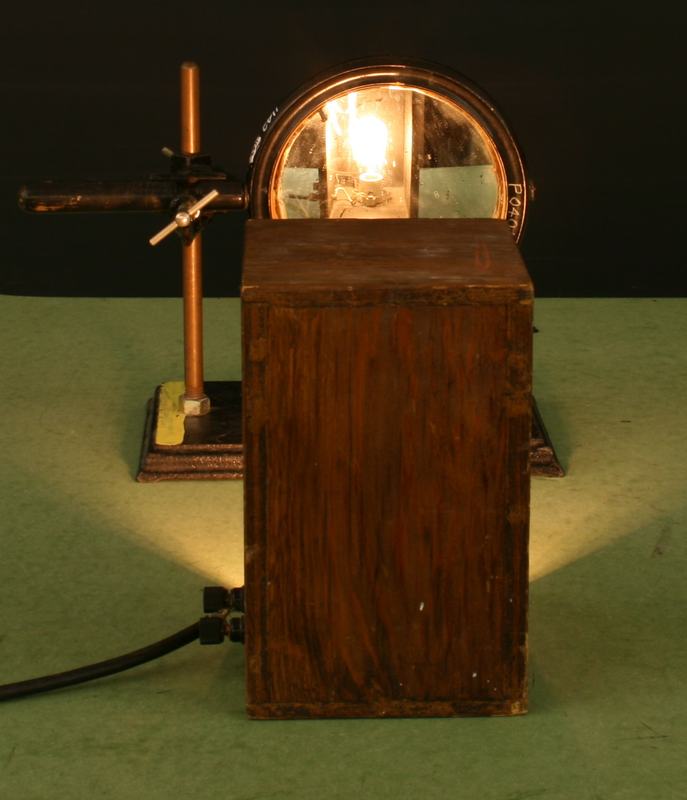 Carbon filament lamp in a wooden shielded box, viewed from the open side with the glowing filament visible; a hand-held mirror on an adjustable stand is positioned behind