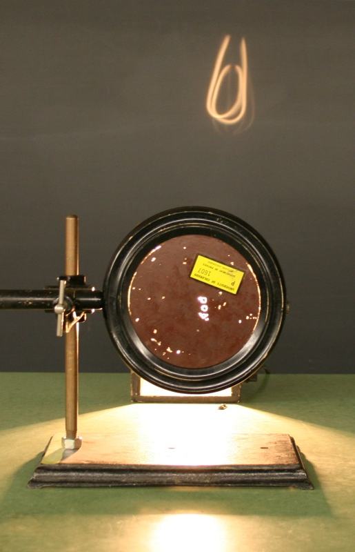 Large concave mirror in a round metal frame on a wooden base, with an adjustable brass lab stand beside it; a projected image of the carbon filament is visible on the blackboard behind
