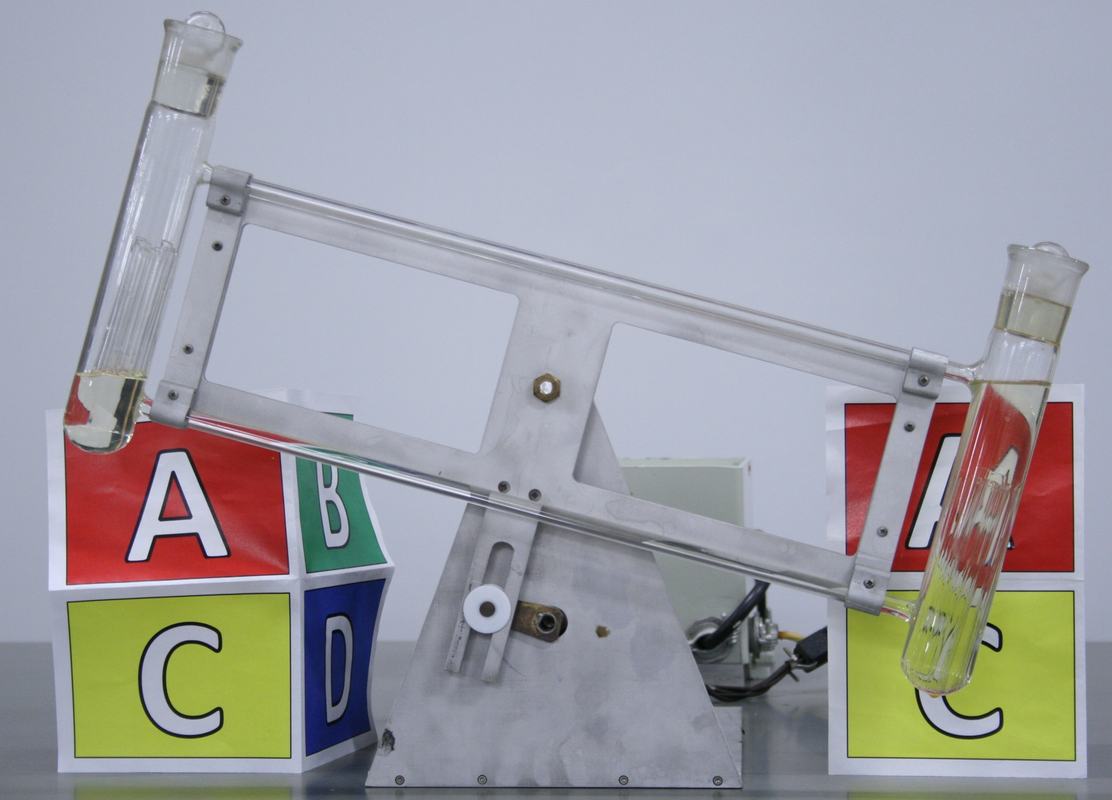 Same tilting frame tilted the opposite direction, now with oil filling the left vial; letter cards visible behind both vials