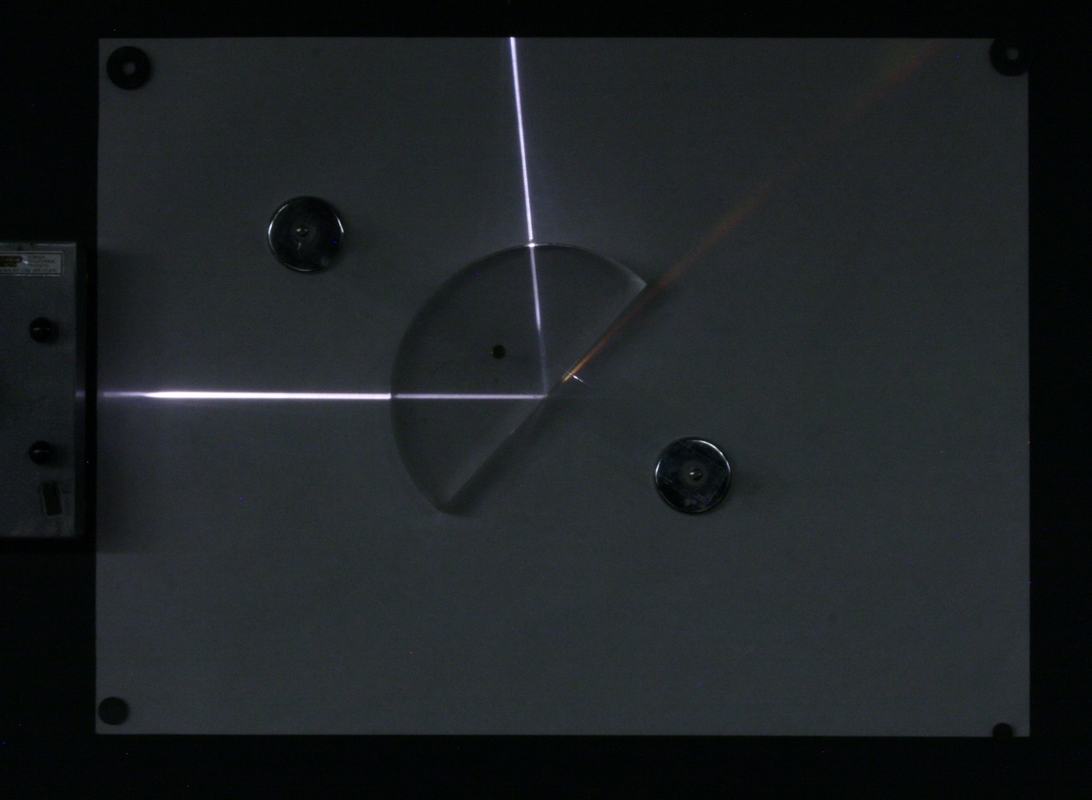 Semi-circular acrylic block on blackboard optics board with a light ray entering the curved surface along a radius, refracting at the flat surface with a partially reflected ray visible