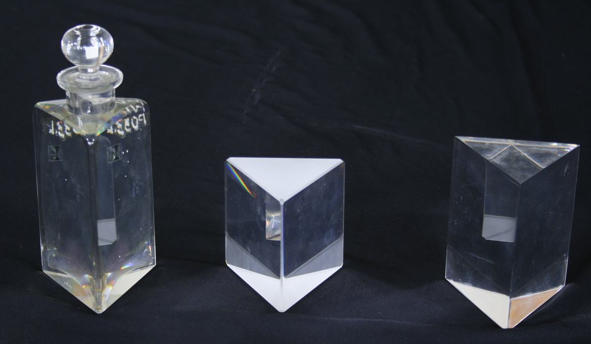 Three prisms on black cloth: glass bottle with stopper containing ethyl cinnamate liquid (left), solid glass prism (center), acrylic prism (right)