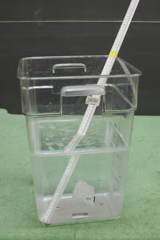 Transparent acrylic container half-filled with water, with a white plastic meter stick placed diagonally showing a visible bend at the water-air interface